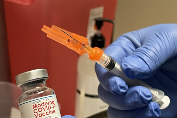 A Moderna vaccine vial and syringe being held by gloved hands.
